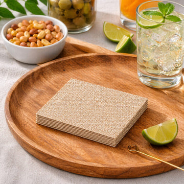 A stack of square woven placemats or coasters in a natural tan jute-like material, displayed on a round wooden serving tray. The scene is styled with a glass of iced drink with lime, a bowl of peanuts, and lime wedges on a light fabric surface.